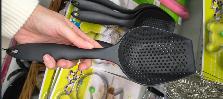Czarna plastikowa łyżka cedzakowa trzymana w dłoni, wyeksponowana nad innymi podobnymi łyżkami na sklepowej półce, w tle widoczne opakowania z grafiką kuchenną.