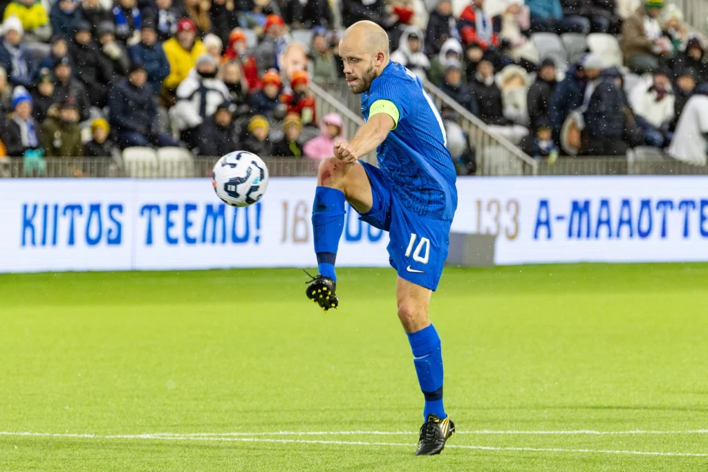 Teemu Pukki w ostatnim meczu w reprezentacji Finlandii Piłkarz w niebieskim stroju wykonuje dynamiczny ruch kopnięcia piłki na murawie stadionu, w tle widownia oraz banery z napisami, a wokół rozproszeni kibice.