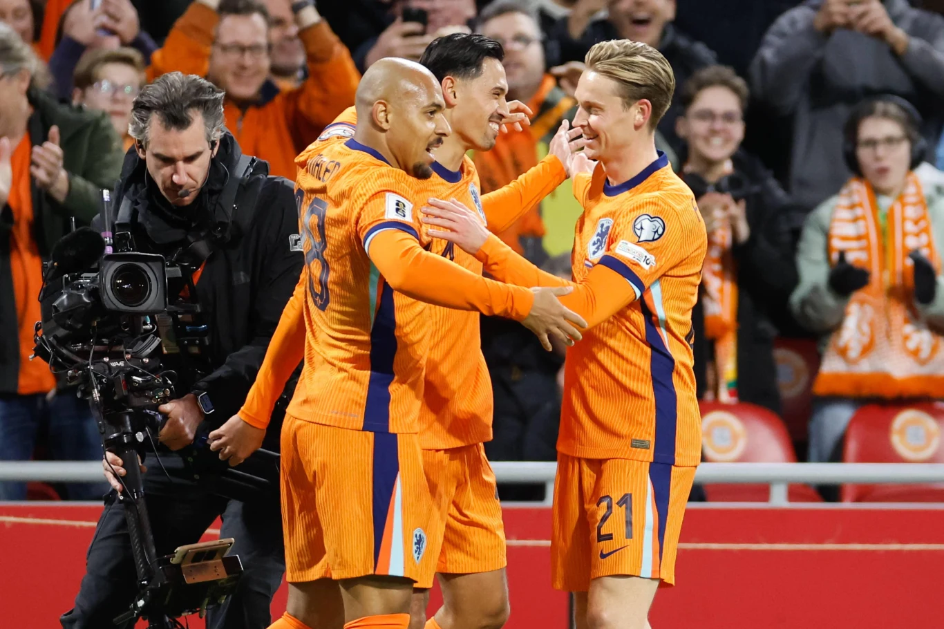 Zawodnicy drużyny piłkarskiej ubrani w pomarańczowe stroje świętują zdobycie bramki na boisku, otoczeni przez zadowolonych kibiców na trybunach i kamerzystę filmującego wydarzenie.