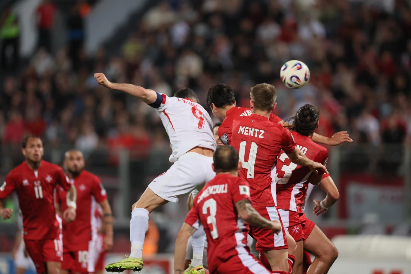 Po tym uderzeniu głową Roberta Lewandowskiego prowadziliśmy w Ta Qali 1:0 Intensywna akcja podczas meczu piłki nożnej, kilku zawodników rywalizuje o piłkę w powietrzu, wszyscy skoncentrowani na zdobyciu przewagi, kibice widoczni w tle tworzą sportową atmosferę.