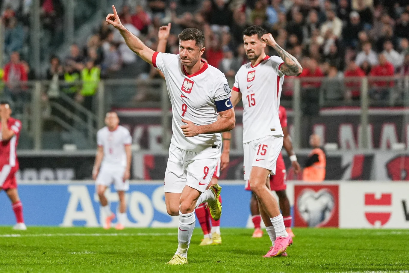 Dwóch piłkarzy reprezentacji Polski w białych strojach świętuje zdobycie gola na tle stadionu pełnego kibiców, jeden z nich unosi rękę w geście triumfu, a drugi stoi tuż za nim z podniesionym palcem