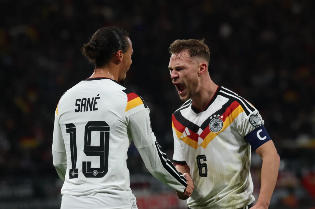 Leroy Sane i Joshua Kimmich Dwóch piłkarzy niemieckiej reprezentacji w trakcie meczu, jeden wyraża silne emocje, krzycząc z zaciśniętą pięścią, drugi stoi tyłem z widocznym nazwiskiem Sané i numerem 19.