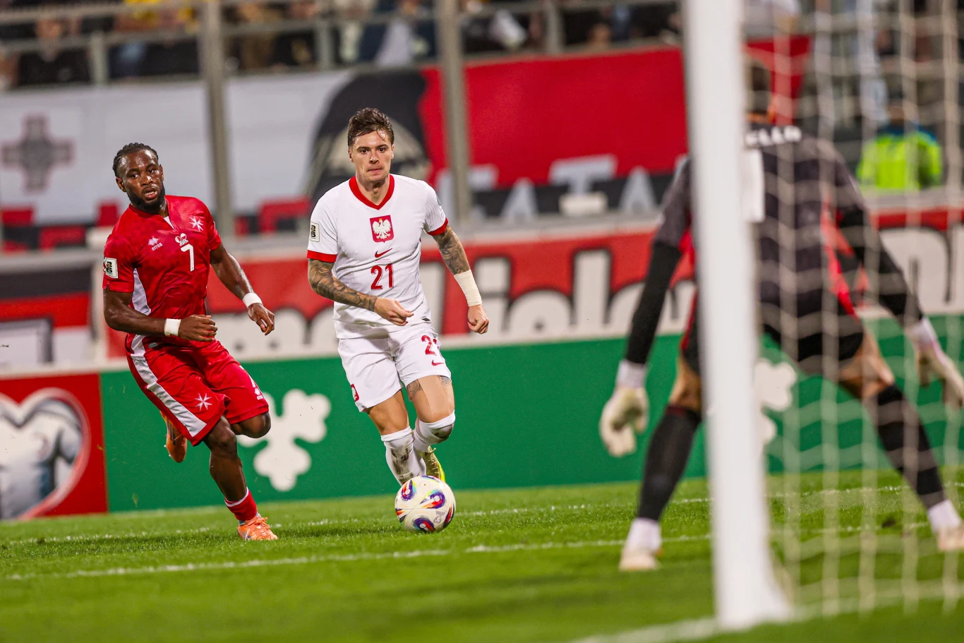 Dwóch piłkarzy reprezentujących przeciwne drużyny ściga się za piłką na boisku piłkarskim. Bramkarz obserwuje sytuację, przygotowując się do ewentualnej interwencji. W tle widoczne są trybuny z kibicami i barierkami reklamowymi.