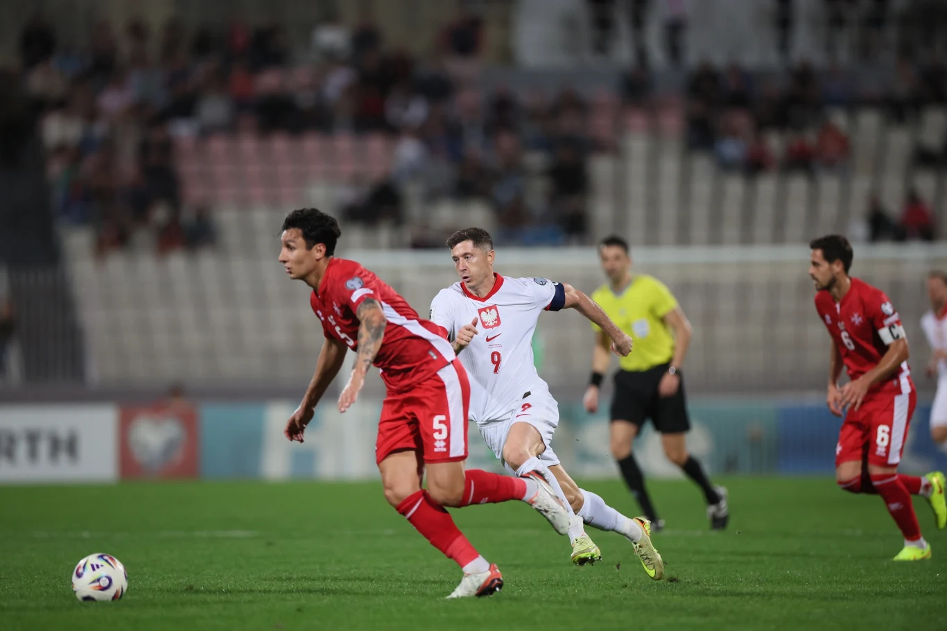Robert Lewandowski goni Kurta Shawa w meczu Malta - Polska Dwóch piłkarzy w dynamicznych pozach walczy o piłkę na murawie, zawodnicy w czerwonych strojach oraz biały piłkarz z numerem 9 są w centrum akcji, sędzia w żółtej koszulce obserwuje przebieg gry, w tle widownia na stadionie.