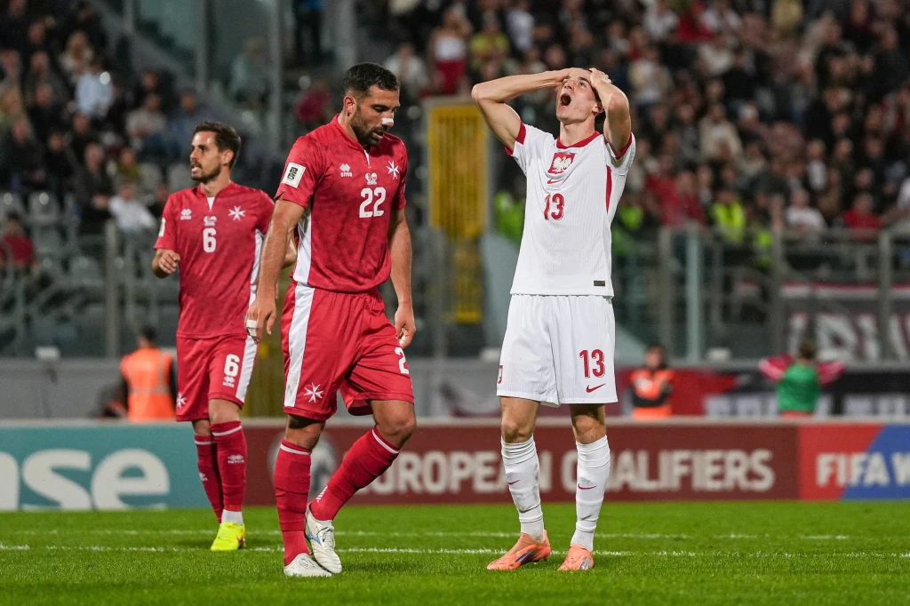 Dwóch piłkarzy drużyny w czerwonych strojach oraz zawodnik w białym stroju numer 13 na środku boiska piłkarskiego. Zawodnik w białym stroju trzyma ręce na głowie w geście rozczarowania, wokół nich widoczni kibice na trybunach.