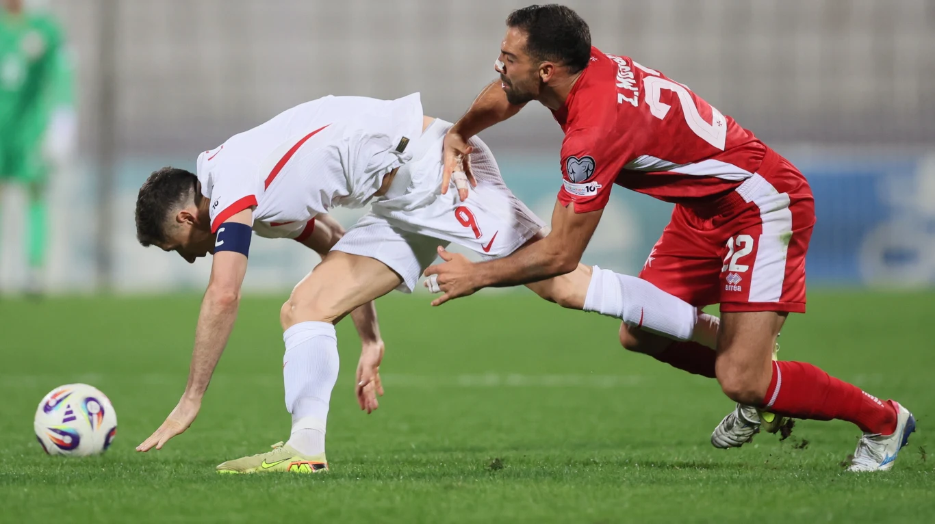 Dwóch piłkarzy rywalizuje na boisku, jeden w białym stroju jest przewracany przez zawodnika w czerwonym, który próbuje zatrzymać przeciwnika poprzez kontakt fizyczny, na murawie piłka do gry.