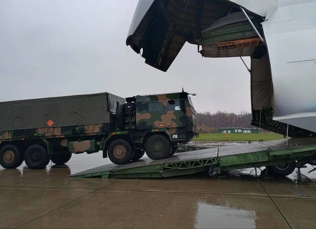 Manewry Northern Strike 225. Polscy żołnierze uczestniczą w ćwiczeniach artyleryjskich w Finlandii Wojskowa ciężarówka w kamuflażu wjeżdża do wnętrza dużego samolotu transportowego, korzystając z metalowej rampy na mokrym lotnisku. W tle widoczne są drzewa i niewielkie budynki.