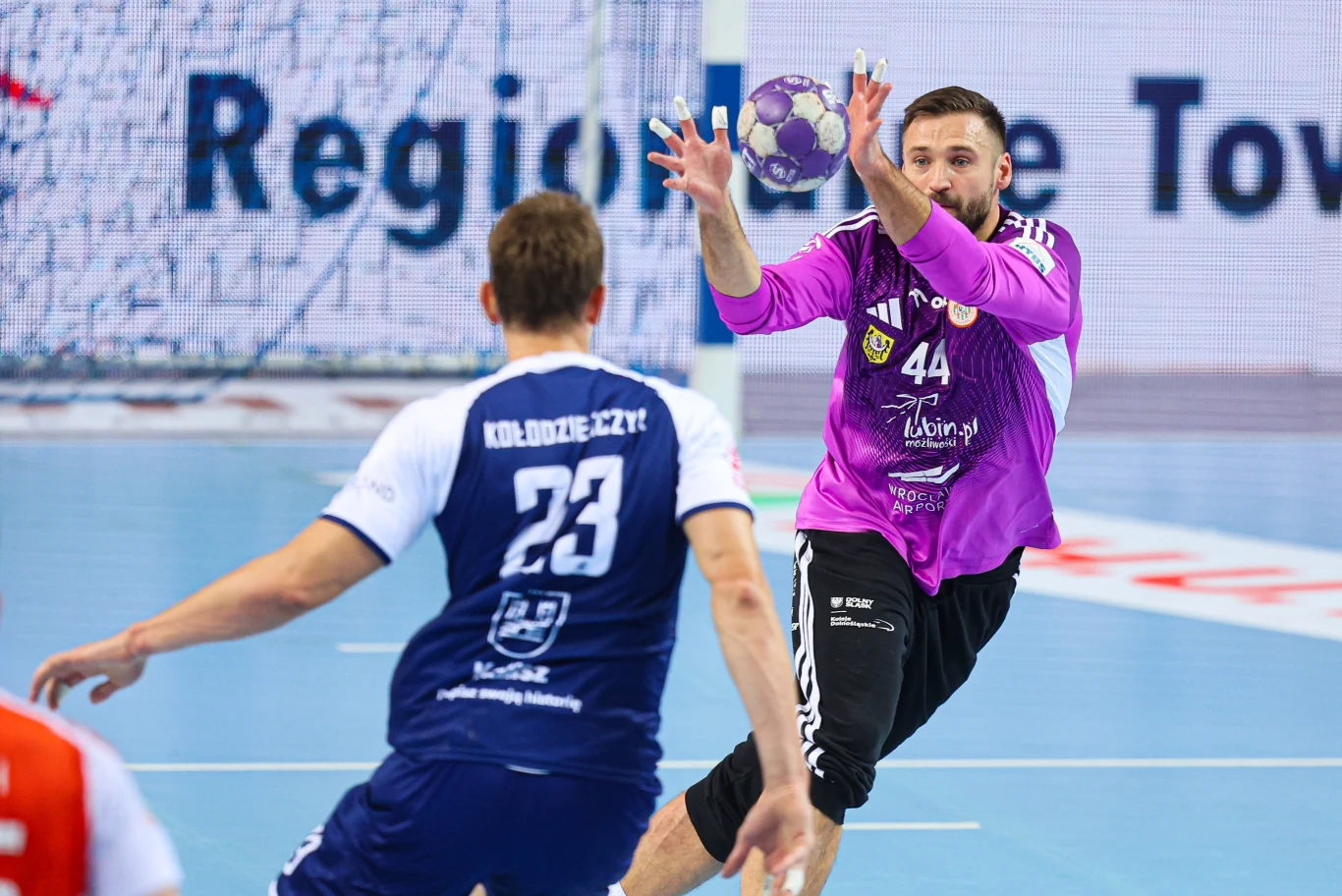 Bramkarz w fioletowym stroju rzuca piłką ręczną, podczas gdy zawodnik drużyny przeciwnej z numerem 23 próbuje zablokować podanie w dynamicznej akcji na hali sportowej.