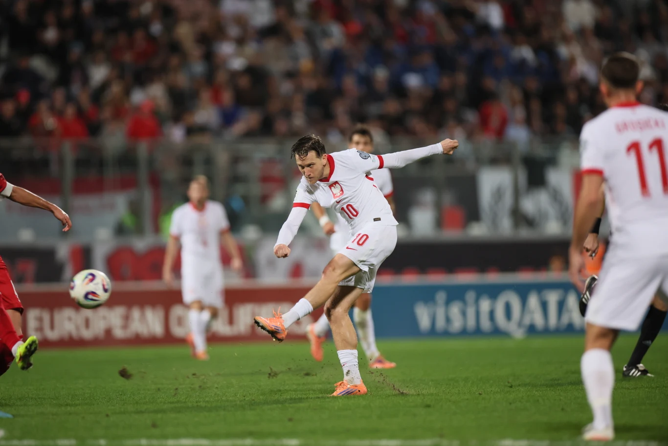 Piotr Zieliński strzelił gola na wagę trzech punktów Zawodnik w białej koszulce z numerem 10 kopie piłkę podczas meczu piłkarskiego, obok na murawie widać innych piłkarzy oraz kibiców na trybunach w tle.