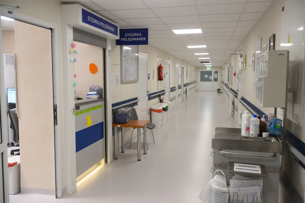 Długi, czysty korytarz szpitalny z otwartymi drzwiami do dyżurki pielęgniarek, wzdłuż ścian znajdują się sprzęty medyczne, tablice informacyjne oraz krzesła, a na końcu korytarza widoczne jest światło.