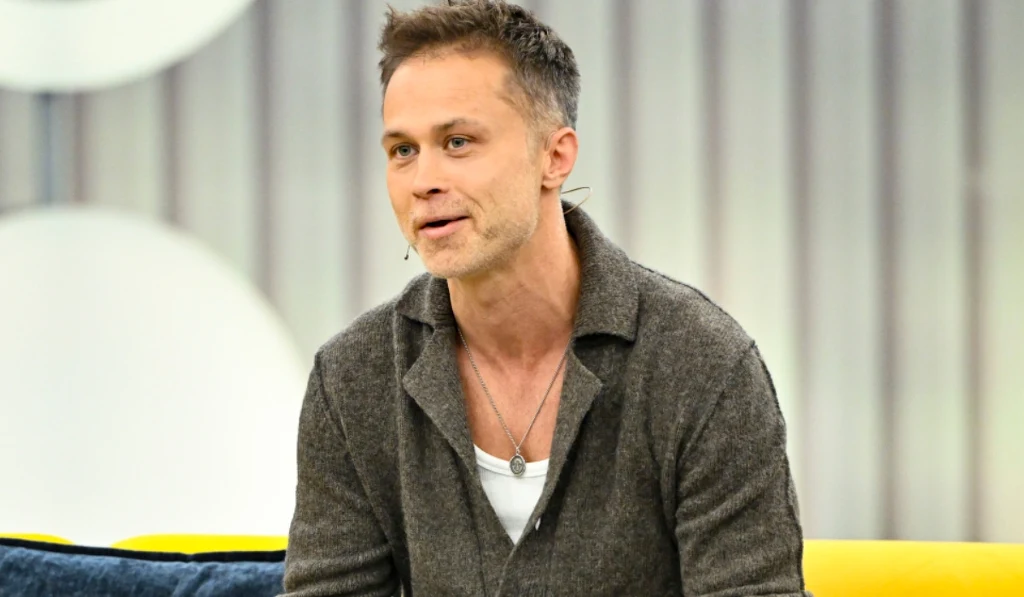 Maciej Zakościelny Mężczyzna w szarej rozpinanej bluzie i białej koszulce siedzi na żółtej kanapie, z mikrofonem przypiętym do ubrania. W tle jasne i szare elementy dekoracyjne.