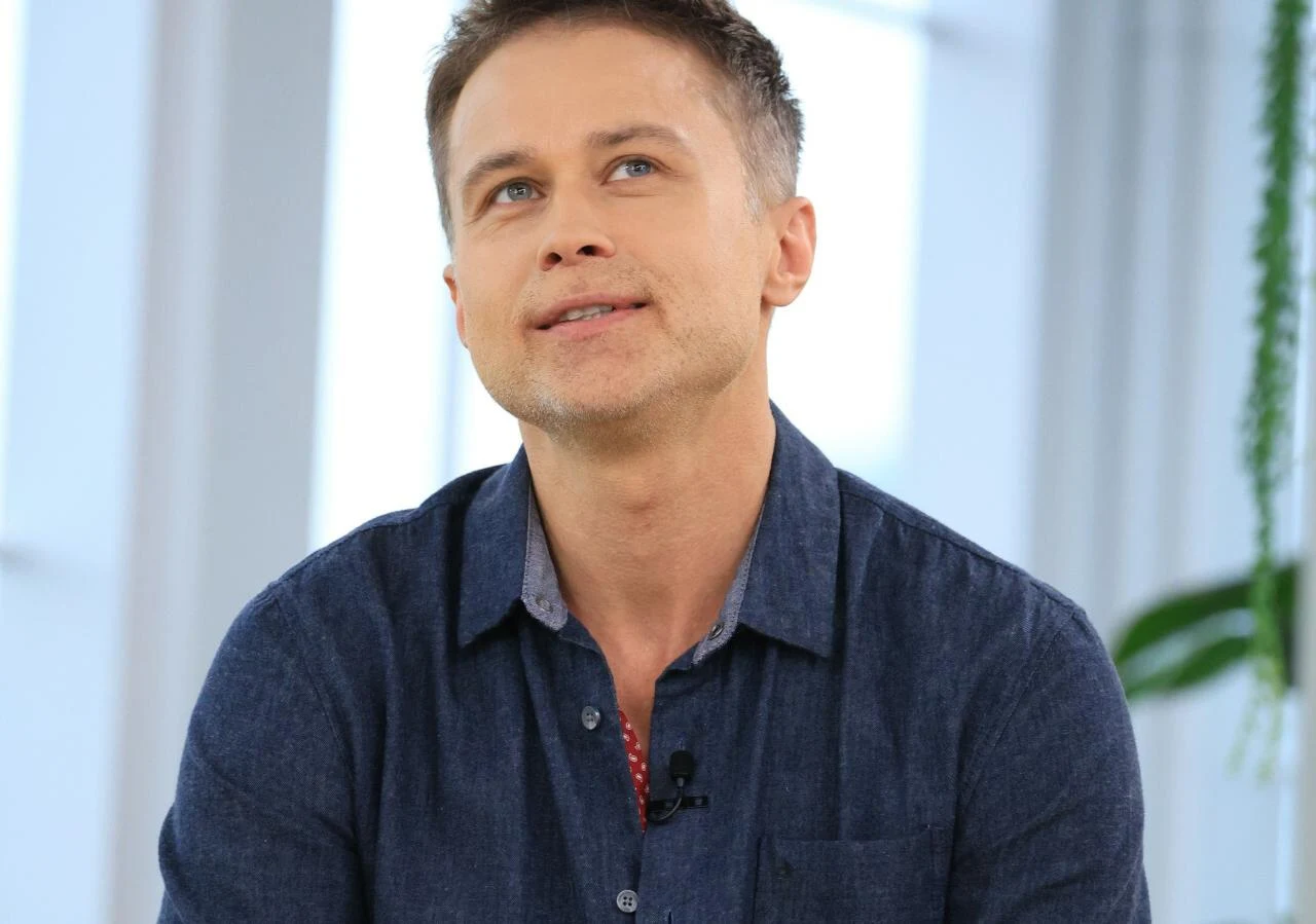 Maciej Zakościelny Mężczyzna w niebieskiej koszuli siedzi w jasnym pomieszczeniu, spogląda lekko w górę, w tle widoczne okno oraz zielona roślina.