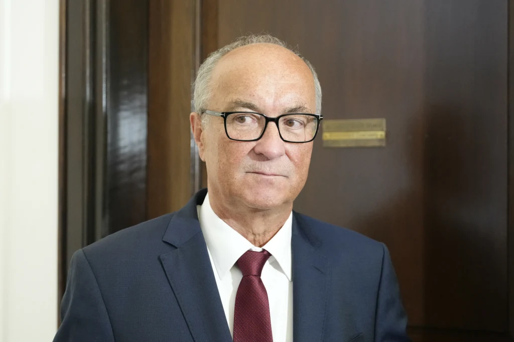 Mężczyzna w średnim wieku w garniturze i bordowym krawacie stoi na tle drewnianych drzwi, na nosie okulary, włosy siwe, wyraz twarzy poważny.