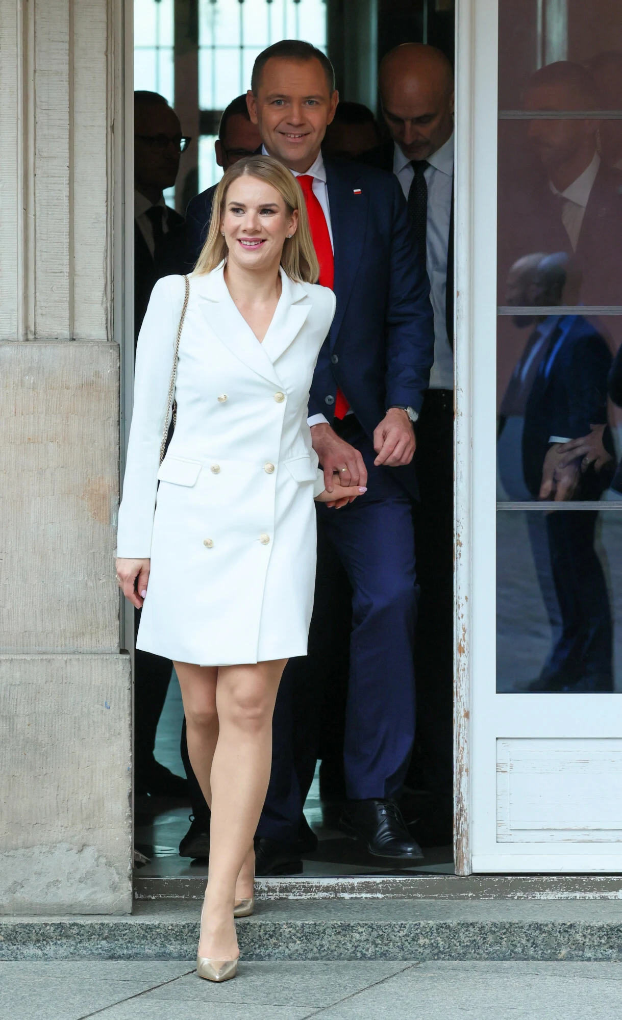 Uśmiechnięta kobieta w białej sukience opuszcza budynek, za nią wychodzą elegancko ubrani mężczyźni w garniturach. Wszyscy wyglądają na uczestników oficjalnego wydarzenia, a atmosfera jest formalna i pozytywna.