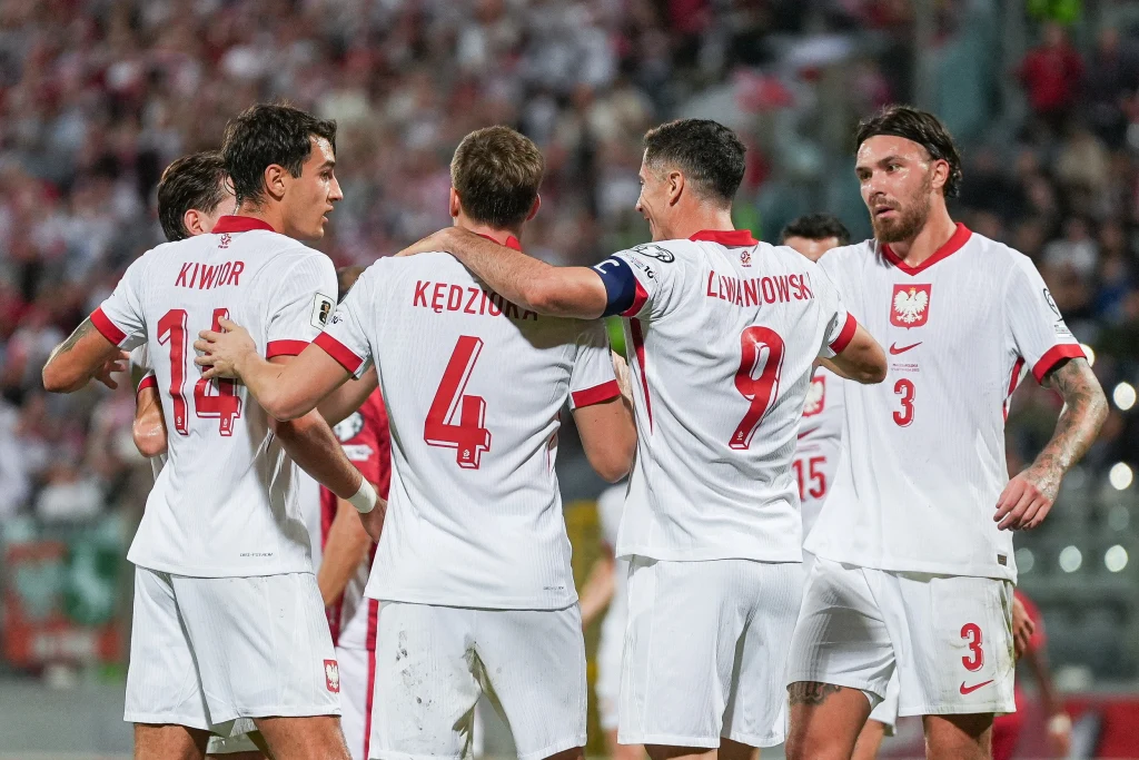 Jakub Kiwior (pierwszy z lewej) Grupa polskich piłkarzy w białych koszulkach z czerwonymi numerami cieszy się wspólnie na boisku, jeden z zawodników obejmuje dwóch kolegów z drużyny, wokół nich kibice na stadionie.