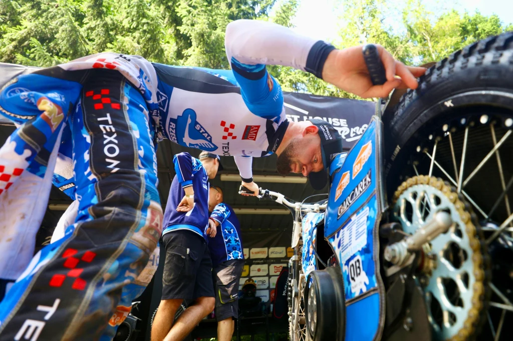 Nicolai Klindt pracuje przy sprzęcie. Zawodnik w stroju żużlowym nachyla się nad motocyklem, sprawdzając stan maszyny. W tle widoczne są inne osoby w zespole oraz otoczenie warsztatu na świeżym powietrzu, dookoła drzewa i sprzęt techniczny.
