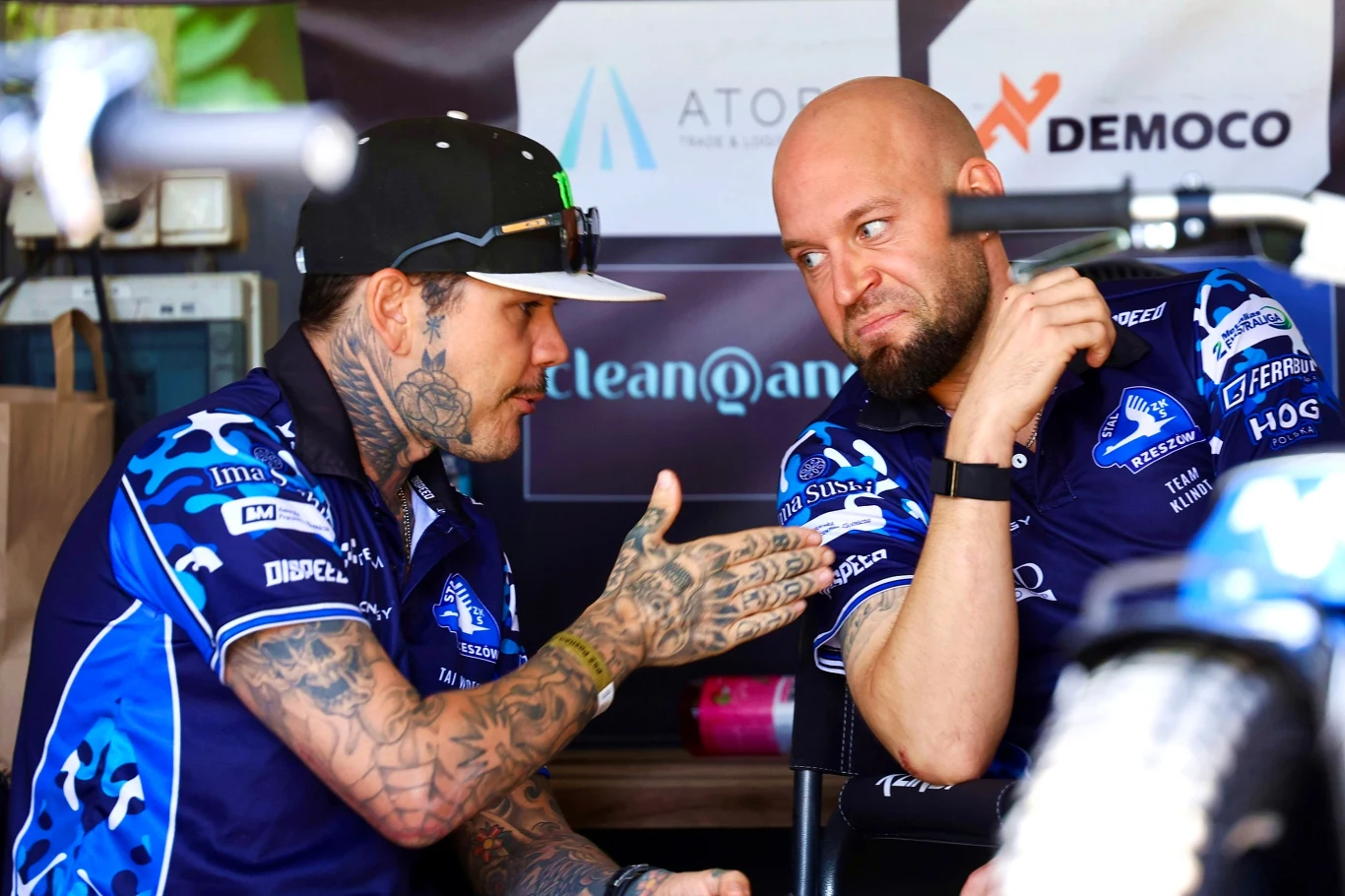 Tai Woffinden w rozmowie ze swoim zmiennikiem Nicolaiem Klindtem. Dwóch mężczyzn w niebieskich koszulkach drużynowych prowadzi rozmowę, jeden z nich ma liczne tatuaże na rękach i czapkę z daszkiem, drugi jest łysy i patrzy z uwagą na rozmówcę. Za nimi znajdują się bannery sponsorów.