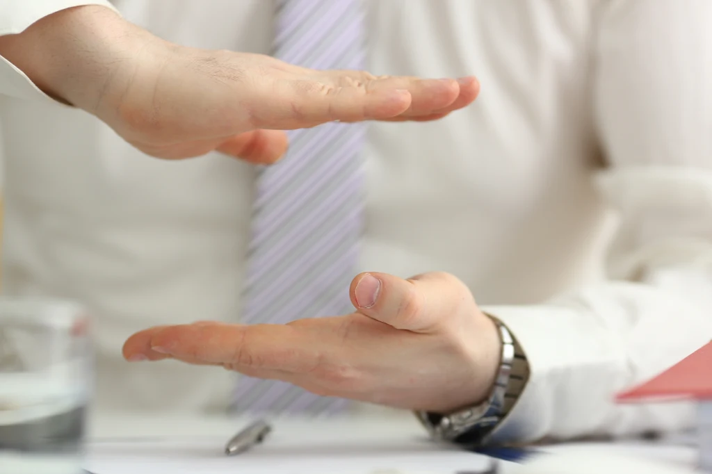 Dlaczego częściej używamy prawej lub lewej ręki? Skąd bierze się dominacja jednej ze stron? Dwie dłonie w geście pokazującym odległość lub wielkość, osoba ubrana w białą koszulę i krawat, na blacie biurka szklanka z wodą oraz długopis.