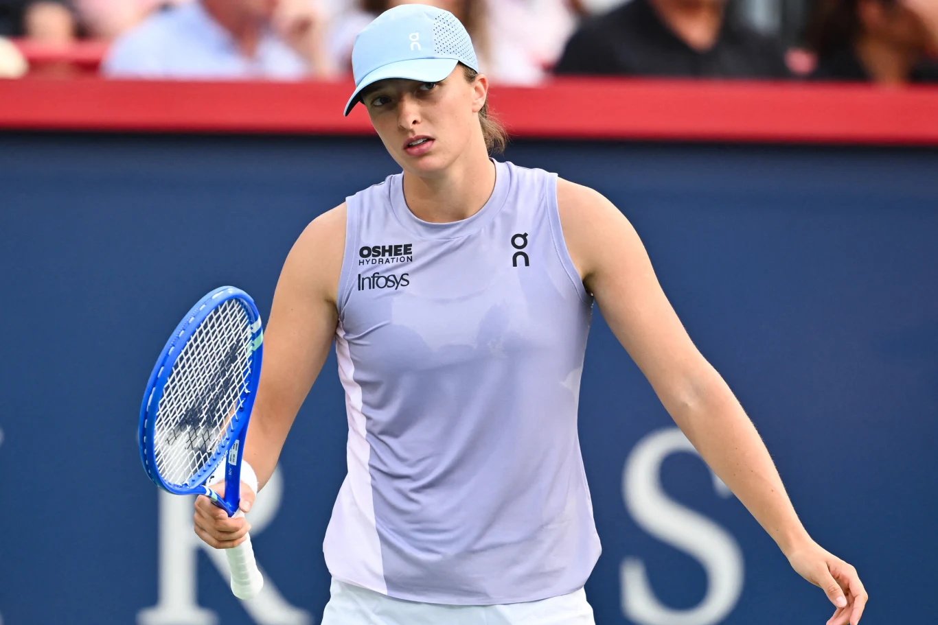 Iga Świątek Tenisistka w sportowej odzieży i jasnej czapce na głowie trzyma rakietę tenisową na korcie, za nią widoczna jest rozmyta publiczność.