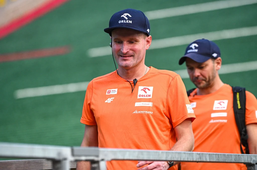 Dwóch mężczyzn ubranych w sportowe, pomarańczowe koszulki i granatowe czapki z logo Orlen, stojących na tle skoczni narciarskiej z zieloną i czerwoną matą.