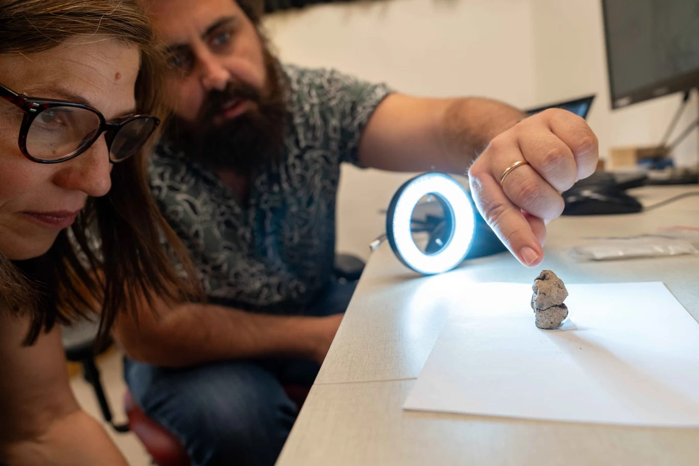 Prehistoryczna figurka sprzed 12 tysięcy lat ujawnia duchowy świat pierwszych osadników nad Jeziorem Galilejskim Dwie osoby skupione na dokładnej obserwacji małego przedmiotu znajdującego się na białej kartce, podświetlonego lampą pierścieniową. Na biurku widoczne są elementy wyposażenia laboratoryjnego i komputer.