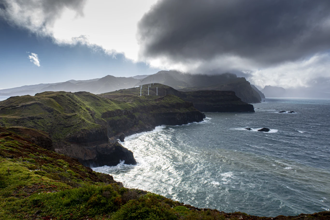 Strome klify porośnięte zieloną roślinnością opadają ku wzburzonemu morzu, nad którym unoszą się ciemne chmury. Po lewej stronie wyraźnie widoczne są białe wiatraki energetyczne, a w tle ciągną się majestatyczne góry.
