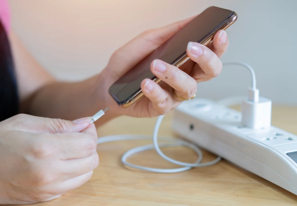 Wiele osób boi się doładowywania telefonu o kilkanaście procent. To błąd! Osoba trzyma smartfon w jednej dłoni, drugą ręką przygotowuje przewód USB do podłączenia do telefonu. Na stole znajduje się listwa zasilająca z kilkoma gniazdkami i podłączoną ładowarką.