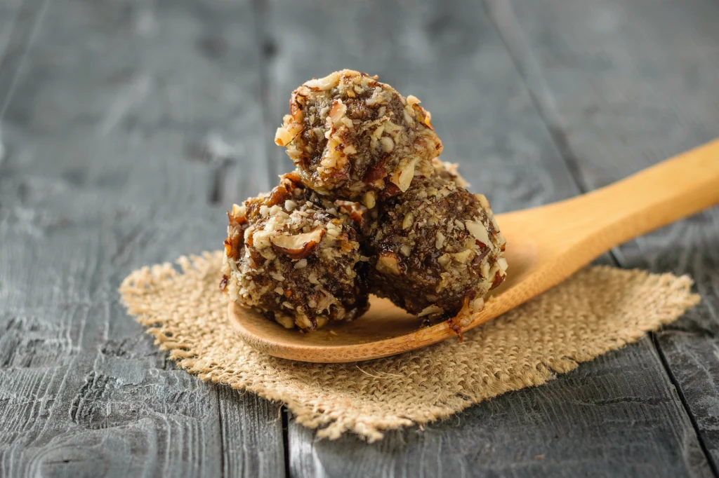 Trzy kulki deserowe obtoczone w pokrojonych orzechach, ułożone na drewnianej łyżce, która spoczywa na jutowej podkładce na tle ciemnego, drewnianego stołu.