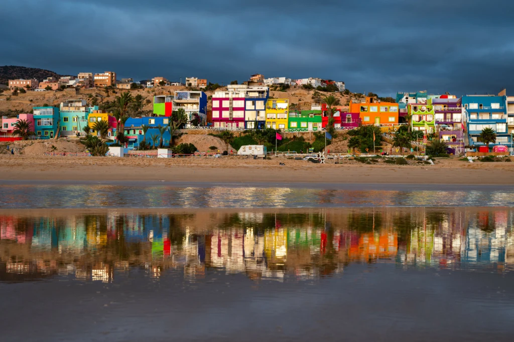 Szereg kolorowych domów stojących na wzgórzu, odbijających się w spokojnej tafli wody u podnóża, z ciemnymi chmurami na niebie kontrastującymi z intensywnymi barwami budynków.