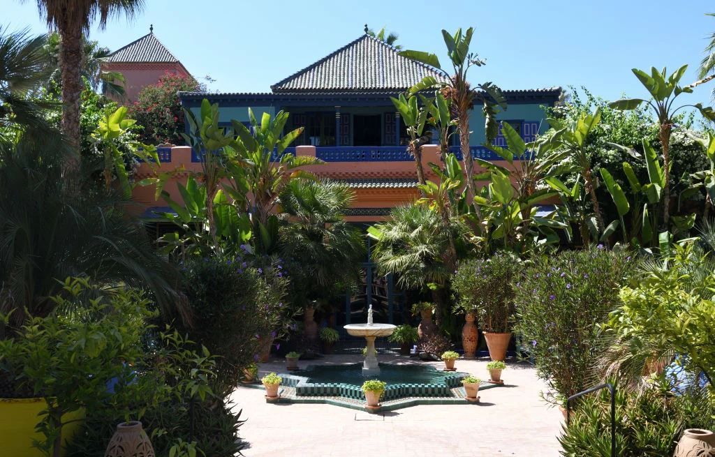 Kolorowy budynek otoczony bujną roślinnością, centralnie usytuowana fontanna otoczona donicami z kwiatami oraz palmami tworzącymi tropikalny klimat ogrodu.
