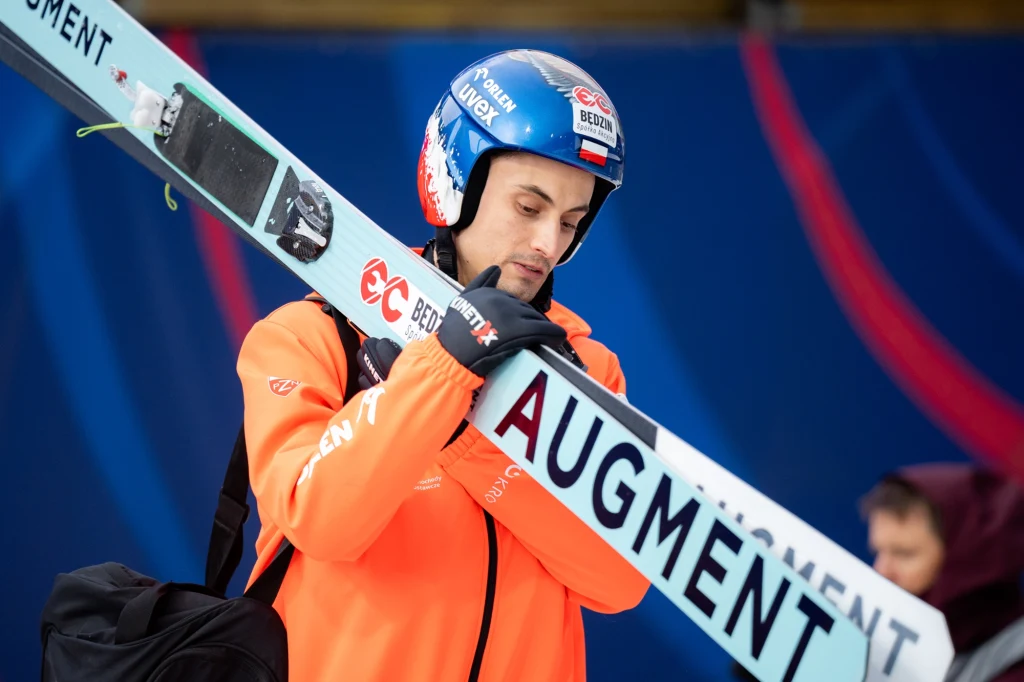 Maciej Kot, 05.04.2025 r. Sportowiec w kasku i pomarańczowej kurtce trzyma narty skokowe z napisem AUGMENT, w tle rozmyta postać oraz niebieskie tło dekorowane czerwonymi elementami.