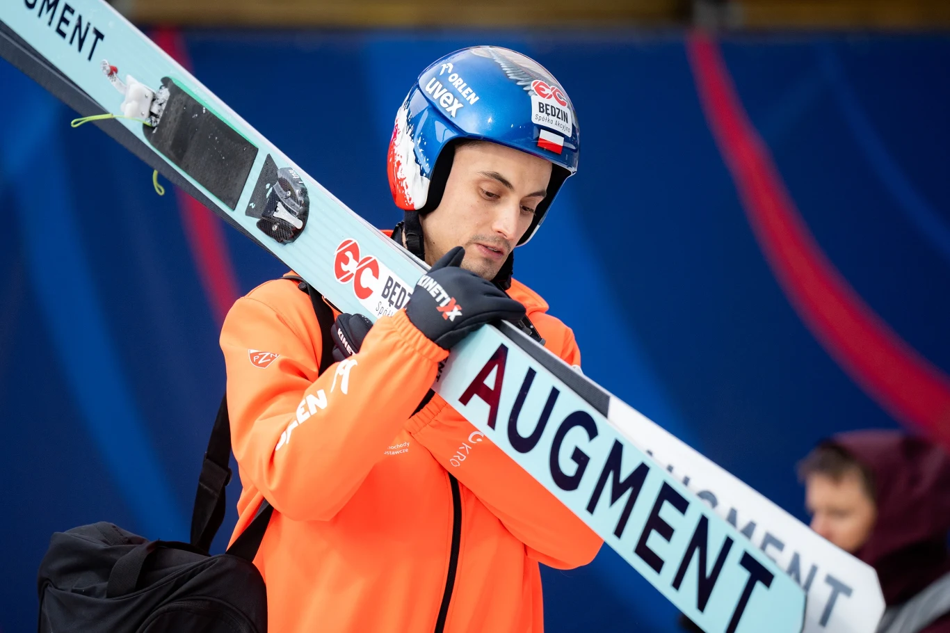 Maciej Kot, 05.04.2025 r. Sportowiec w kasku i pomarańczowej kurtce trzyma narty skokowe z napisem AUGMENT, w tle rozmyta postać oraz niebieskie tło dekorowane czerwonymi elementami.