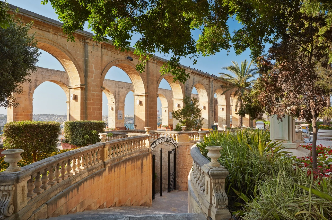Kamienne arkady akweduktu z balustradą schodzącą do ogrodu, otoczone bujną roślinnością, palmami i krzewami, prześwitujące jasne niebo w tle.