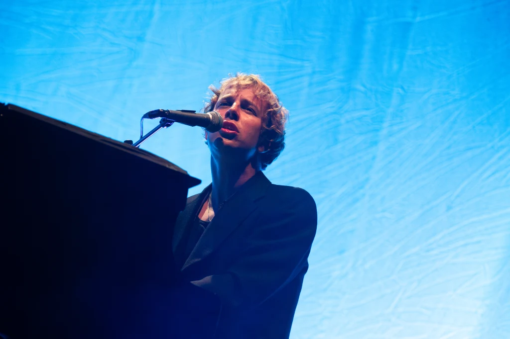 Muzyk z jasnymi, kręconymi włosami wykonuje utwór na scenie, grając na fortepianie i śpiewając do mikrofonu, w tle jednolite, niebieskie oświetlenie.