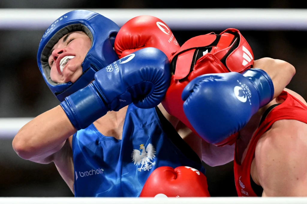 Aneta Rygielska (z lewej) Pojedynek bokserski, w którym zawodniczka w niebieskim kasku otrzymuje silny cios od przeciwniczki w czerwonym stroju. W tle widoczny ring z linami.