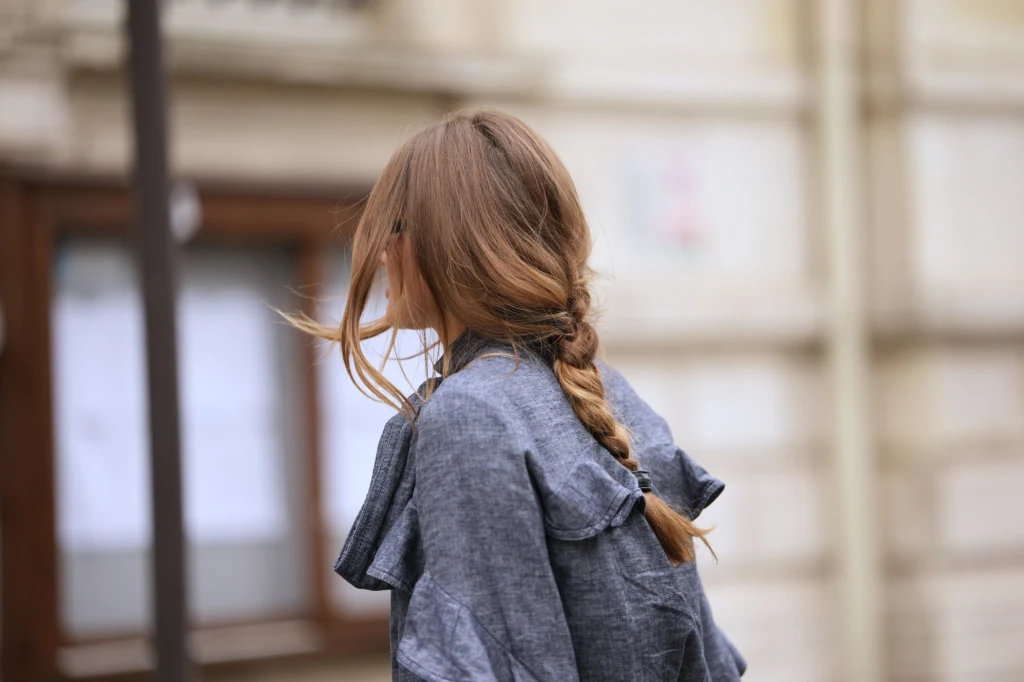 Modne fryzury dla leniwych Kobieta z długimi, blond włosami splecionymi w warkocz ubrana w szarą kurtkę stoi tyłem na tle rozmytego budynku miejskiego.