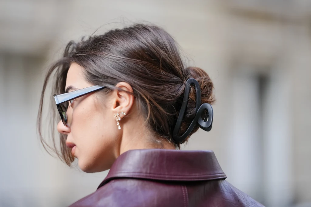 Proste i szybkie fryzury dla kobiet Kobieta z upiętymi włosami ozdobionymi dużą klamrą, ubrana w bordową skórzaną kurtkę oraz nosząca czarne okulary przeciwsłoneczne, widoczna z profilu na tle rozmytego tła miejskiego.