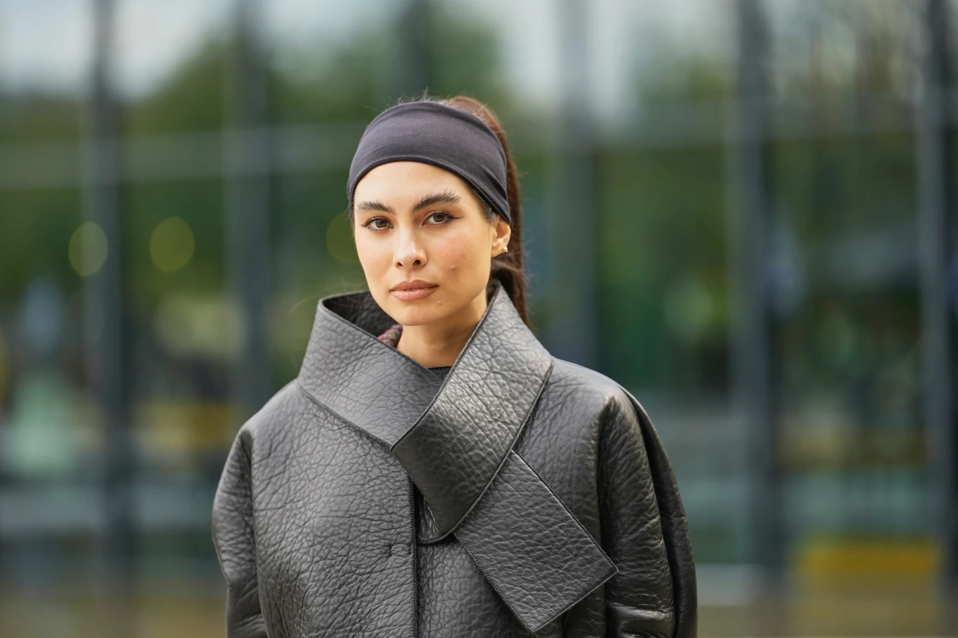 Młoda kobieta z ciemnymi włosami spiętymi opaską ubrana w nowoczesny czarny płaszcz o fakturze skóry, stoi na tle przeszklonego budynku, patrząc poważnie w stronę obiektywu.