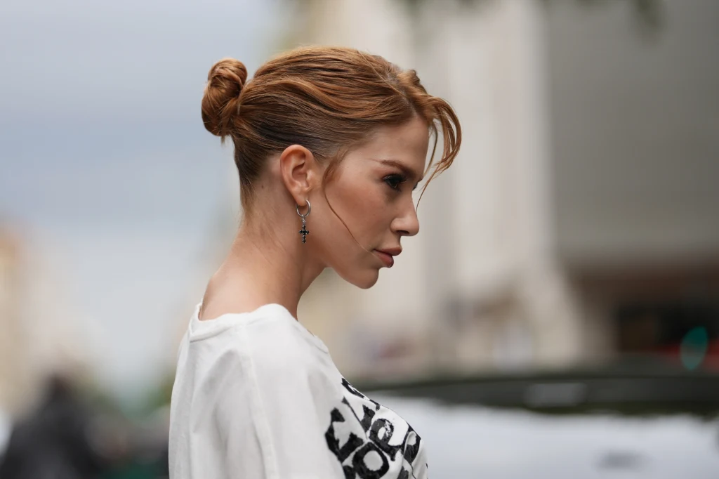 Jak zrobić idealny messy bun? Profil kobiety z upiętymi włosami i delikatnym makijażem, ubranej w jasną bluzkę z czarnym nadrukiem, na tle rozmytego miejskiego krajobrazu.