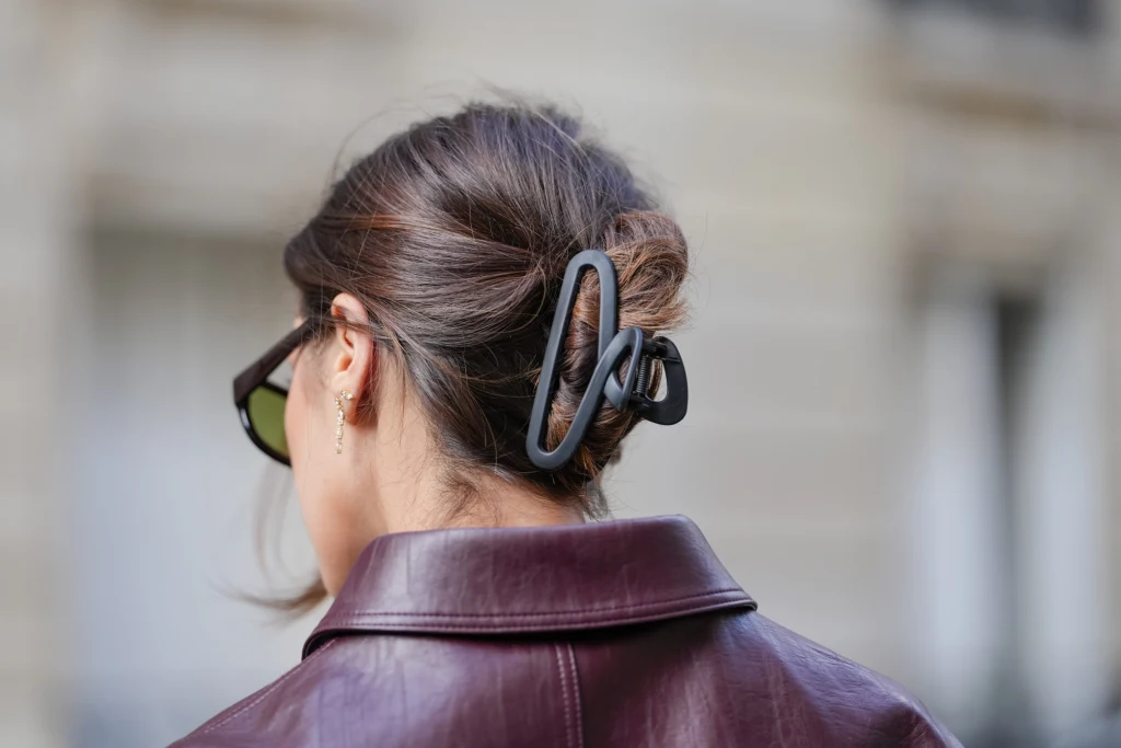 Modne fryzury na zimę Kobieta w ciemnych okularach i bordowej kurtce z włosami spiętymi czarną klamrą, uchwycona z tyłu.