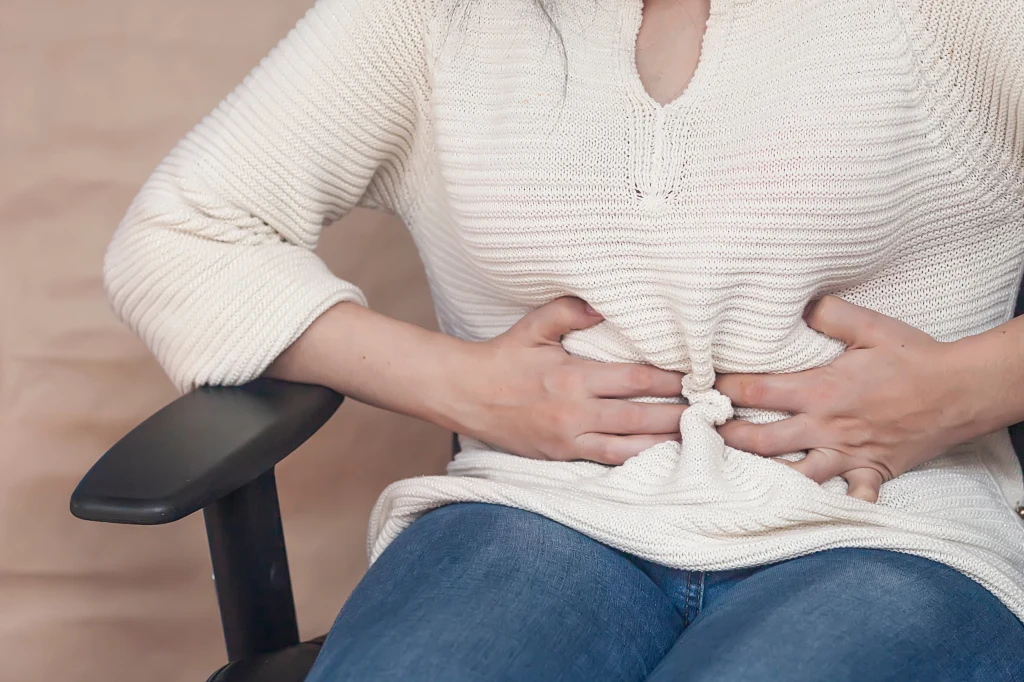 Osoba siedzi na krześle, trzymając ręce na brzuchu, sprawiając wrażenie odczuwania bólu lub dyskomfortu. Ubrana w jasny sweter i dżinsy, widoczna jest tylko górna część ciała bez pokazania twarzy.