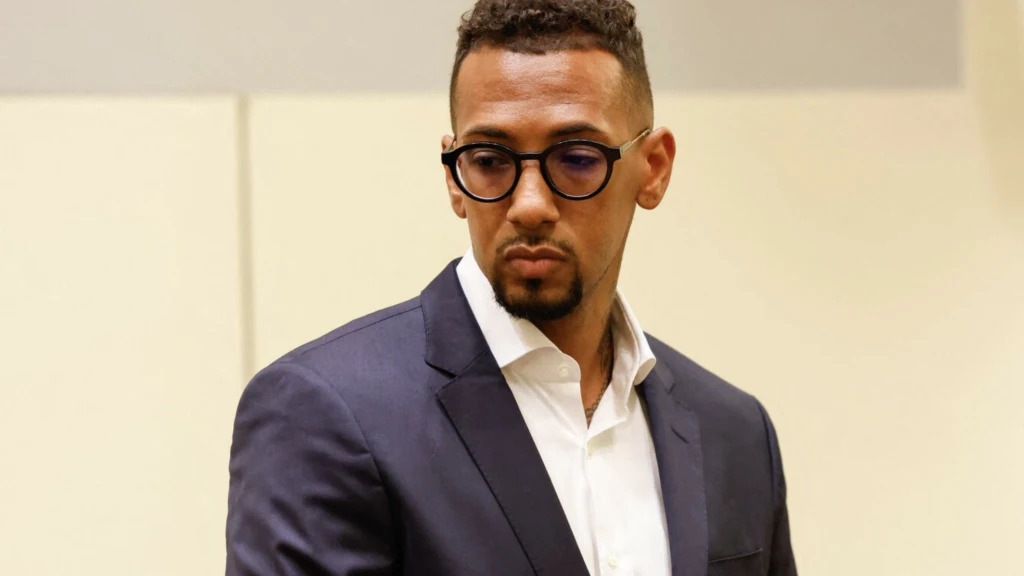 Jerome Boateng Mężczyzna w eleganckiej, ciemnej marynarce i białej koszuli, noszący okulary o ciemnych oprawkach, ma krótkie, kręcone włosy i starannie przyciętą brodę, stoi na neutralnym tle o jasnych barwach.