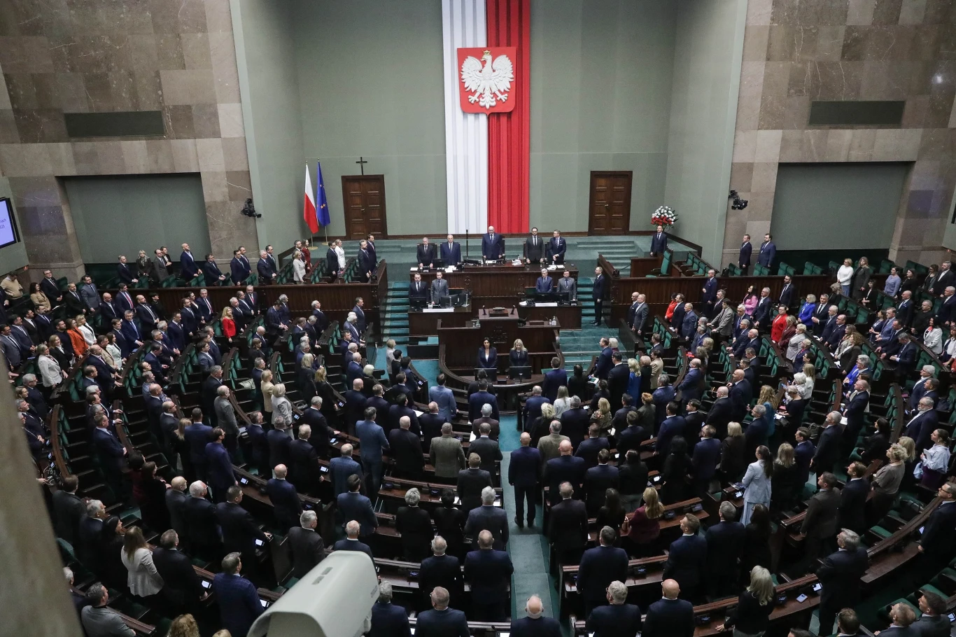 Sala posiedzeń polskiego parlamentu wypełniona posłami, widoczne centralne miejsce prezydialne, duży biało-czerwony sztandar z godłem Polski oraz flagi państwowe po lewej stronie. Wszyscy zgromadzeni stoją, liczna obecność osób wskazuje na ważne wydarz...