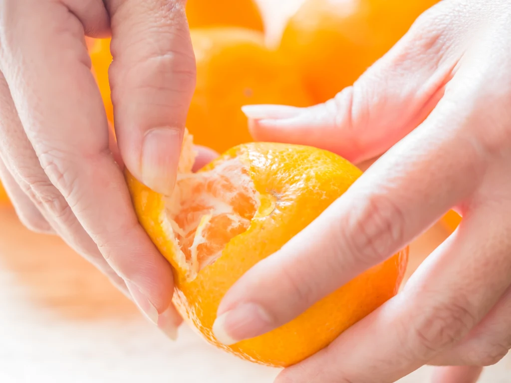 Łatwość obierania mandarynek jest jednym z kryteriów, na których nam zależy Ręce obierające dojrzałą mandarynkę, w tle widoczne rozmyte pomarańczowe owoce.
