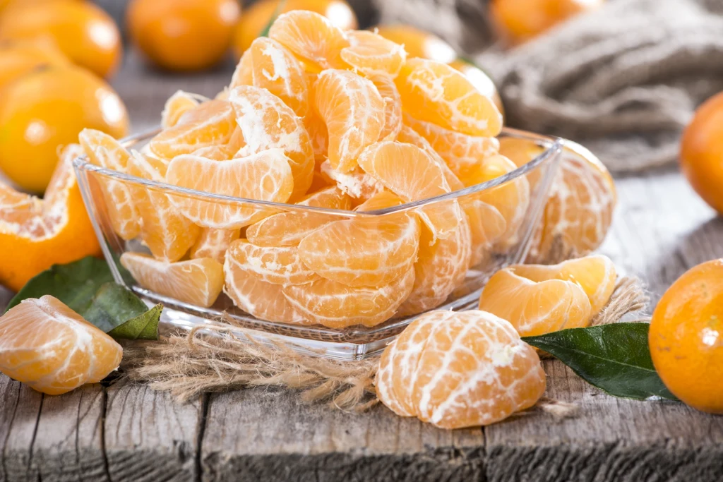 Gdy zostanie nam nadmiar mandarynek, łatwo wykorzystamy je w koktajlach, sałatkach czy jako dodatek do ciast Soczyste cząstki mandarynek ułożone w szklanej misce na drewnianym stole, wokół znajdują się całe, nieobrane mandarynki oraz zielone liście.