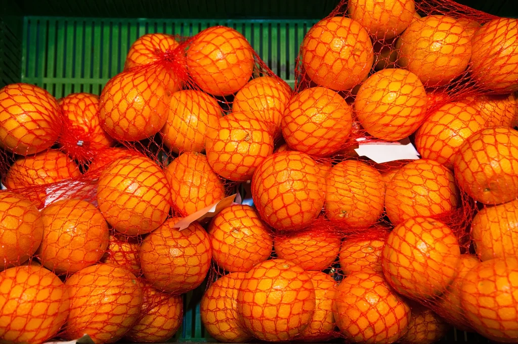 Wiele osób wybiera siatkowane mandarynki, ale jeśli chcemy być pewni, lepiej kupujmy te sprzedawane luzem Pomarańcze zapakowane w pomarańczowe siatki ułożone na zielonej powierzchni, dominują intensywne barwy owoców.