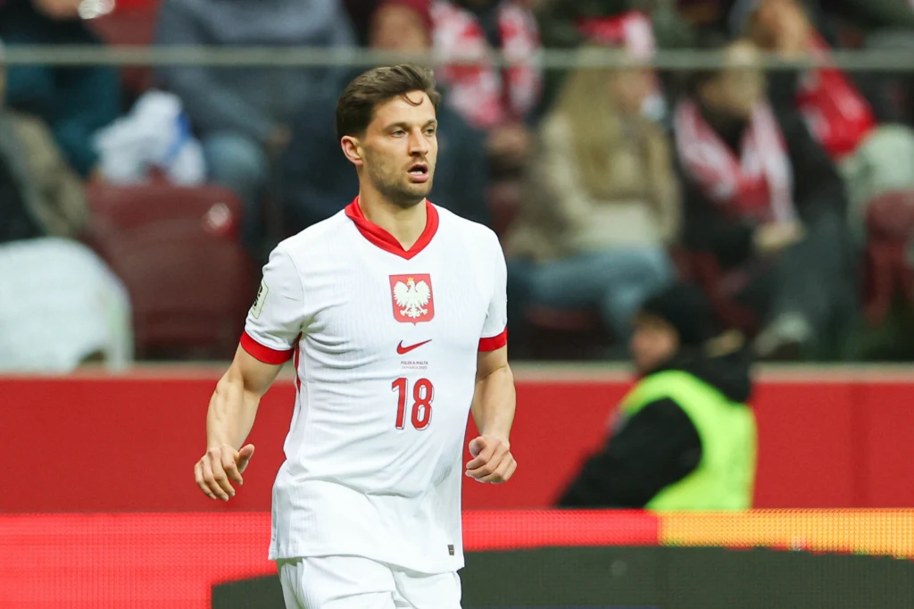 Piłkarz w białej koszulce reprezentacji Polski z numerem 18 na piersi biegnie po boisku, w tle widoczni kibice na trybunach.