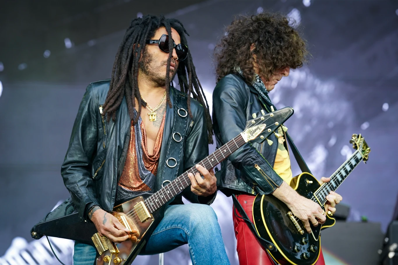 Lenny Kravitz i jego muzyczny partner Craig Ross Dwóch gitarzystów na scenie koncertowej, ubranych w skórzane kurtki i okulary przeciwsłoneczne, grających na elektrycznych gitarach. W tle widoczna scena z reflektorami. Klimat dynamiczny, energetyczny, podkreślający pasję do muzyki rockowej.