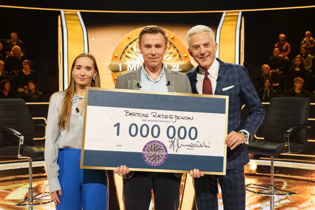 Gra może być inwestycją. Sposoby na „Milionerów” Trzy osoby stoją na scenie programu telewizyjnego, trzymając duży, symboliczny czek na sumę miliona złotych; w tle widoczna publiczność i charakterystyczne elementy scenografii programu quizowego.