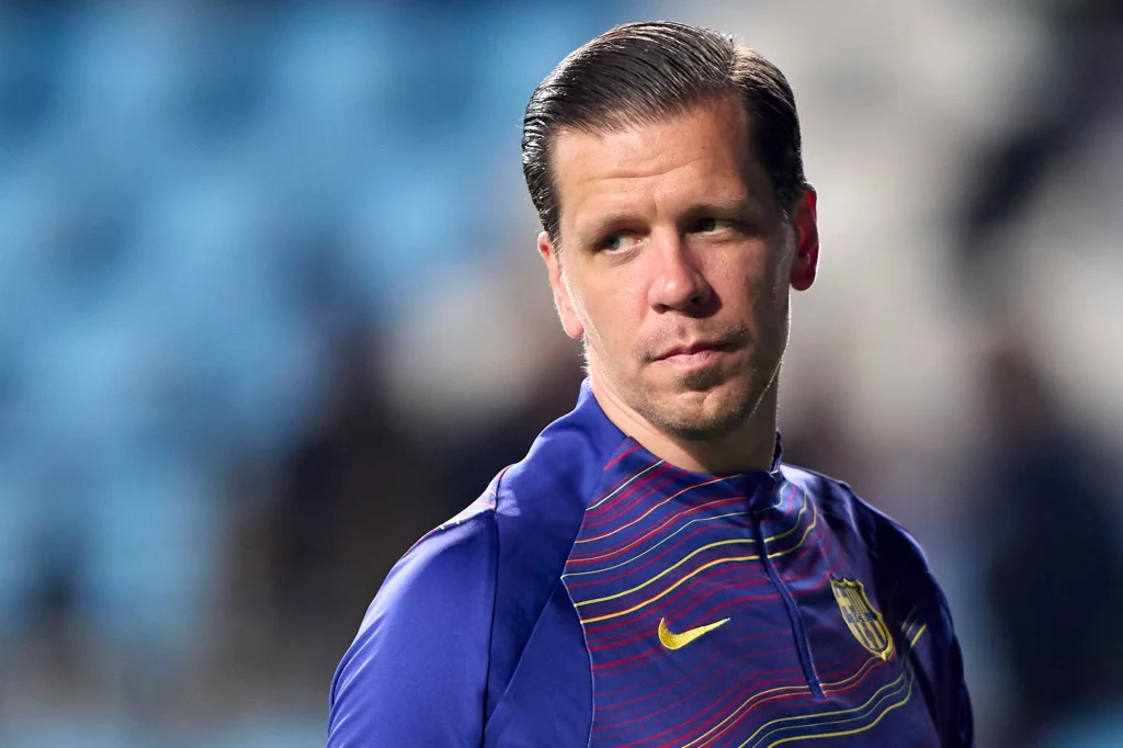Wojciech Szczęsny Mężczyzna w sportowej koszulce z herbem FC Barcelona, patrzący w bok na tle rozmytych, niebieskich miejsc na trybunach stadionu.